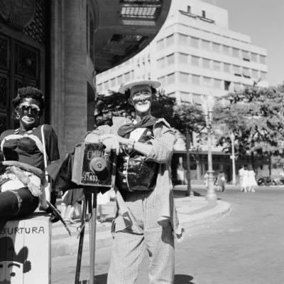 Carnaval de rua, homens fantasiados de mulher e de fotógrafo lambe-lambe