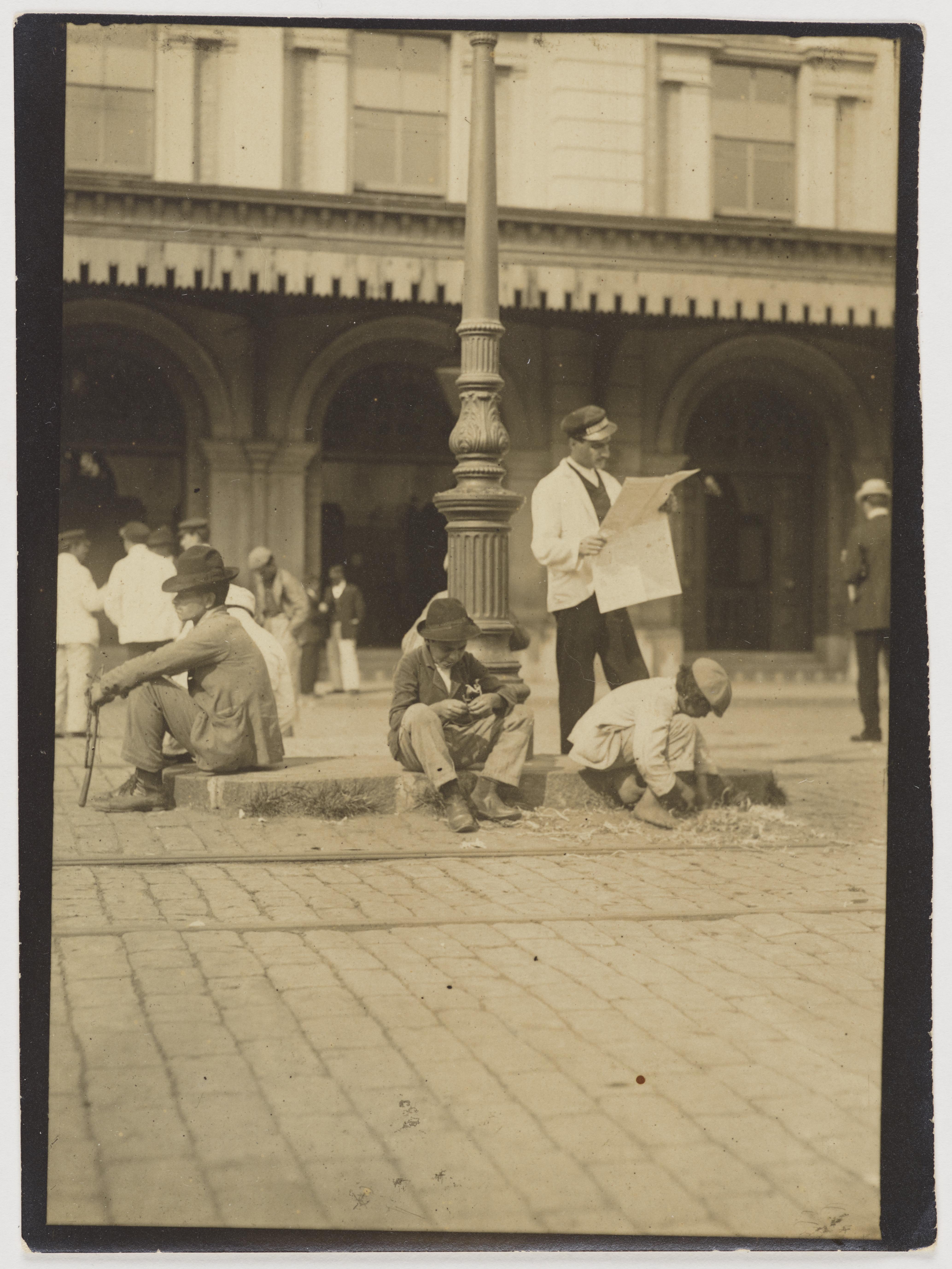 Ferroviário lê jornal, cercado por carregadores de malas,